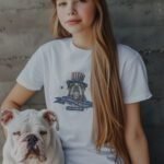 A girl with long hair wearing an Independence Day 4th July | ALL AMERICAN BULLDOG | Unisex Heavy Cotton Tee, featuring an illustration of a bulldog in a patriotic hat, sits next to an American Bulldog.