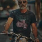 A mature man with a beard riding a motorcycle on a sunny street, wearing sunglasses and a Ride Forever in Style with the Forever 2 Wheel Motorcycle Unisex T-shirt - Your Passport to Timeless Co.