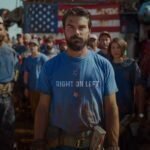 A man with a beard, wearing a Democrat Right on Left | Election 2024 | Unisex Heavy Cotton Tee, stands in front of a crowd at an outdoor rally, looking calmly at the camera.