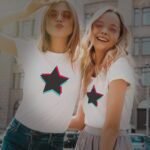 Two smiling young women in sunglasses posing playfully on a sunny city street, both wearing white t-shirts with black Unisex TikTok Star designs.