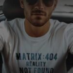 Man in sunglasses and a T-shirt emblazoned with the humorous pun "matrix:404 reality not found" sitting in a vehicle.