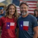 Two people, one in a red Star Flag | Yeah America 24 | Unisex t-shirt and the other in a blue one, both displaying the Texas star flag, are standing outdoors with an American flag in the background.