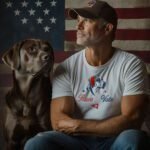 A man in a baseball cap sits with a large dog in front of an American flag, proudly wearing the "Paws to Vote | Elections 2024 | Unisex t-shirt" adorned with an illustration of a dog and stars, making a statement for the upcoming elections.